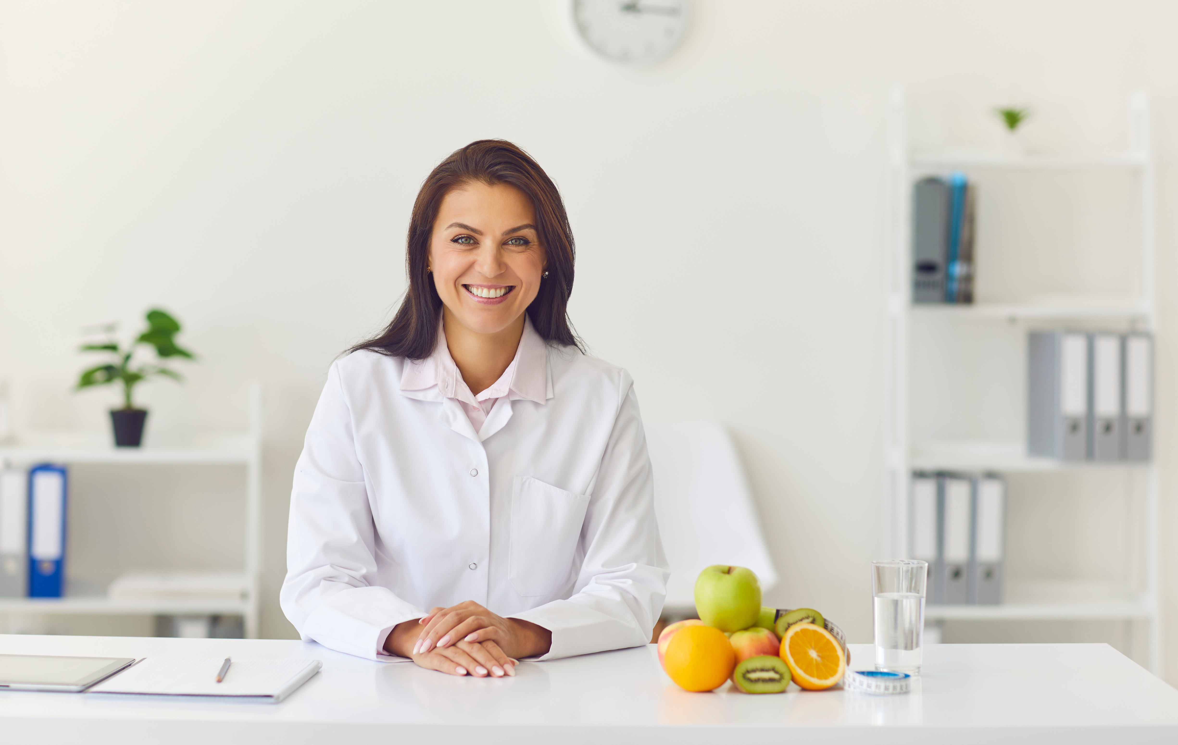 The image shows a doctor with a warm smile sitting at a table. On the table, there are various fruits, a glass of water, and a measuring tape. The presence of these items signifies the focus on healthy eating, hydration, and tracking progress in weight loss at the hormone replacement therapy clinic.